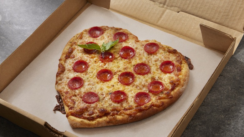 A heart-shaped pepperoni pizza with fresh basil sprig in a takeout box