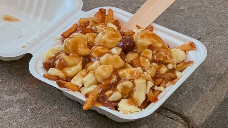 A serving of poutine in a white takeout container, featuring golden fries topped with cheese curds and brown gravy, placed on a concrete surface