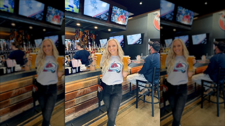 A smiling young blonde woman holding two large drafts of beer inside a bar filled with TVs