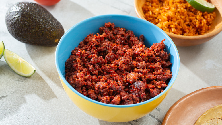 A bowl of ground Mexican chorizo on a table next to a wedge of lime, a whole avocado, and a bowl of rice.