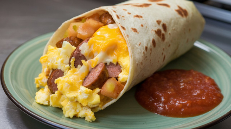 Breakfast burrito filled with egg, cheese, sausage, and potatoes on a plate next to some salsa.