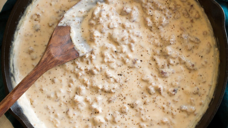 A wooden spoon in a cast iron pan filled with creamy sausage gravy.