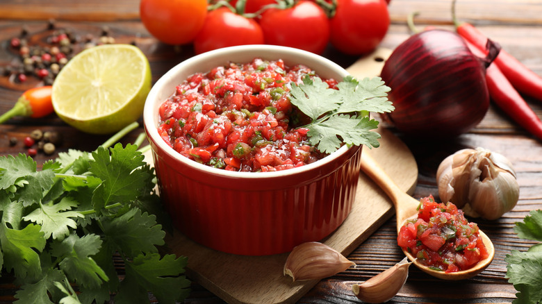 A red ramiken filled with salsa on a wooden table surrounded by fresh onion, lime, garlic, and cilantro.
