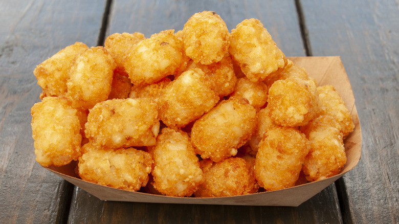 A paper boat filled with crispy tater tots on a wooden table.