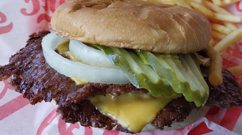 closeup of freddy's smash burger with onions, cheese, and pickles wrapped in paper next to fries