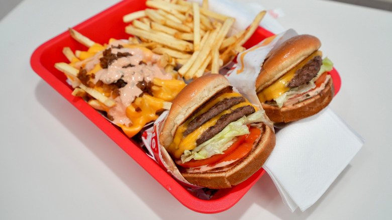 two in n out burgers side by side on a red tray with cheese, lettuce, tomatoes, and sauce with two orders of fries behind them, one with sauce, cheese, and grilled meat