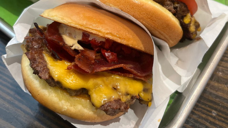 shake shack burger wrapped in white paper with cheese, bacon, and sauce on a silver tray on a wooden table