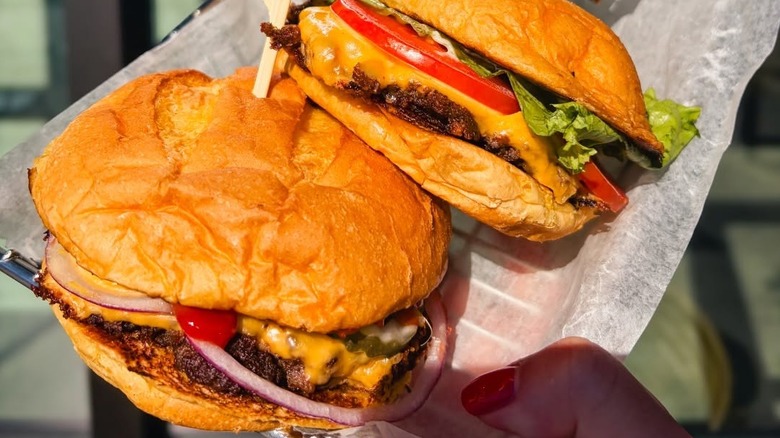two smashburger burgers with cheese and toppings behind held up by a woman's hand on white paper