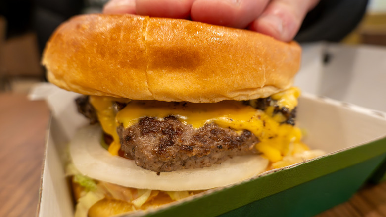 closeup of a wahlburger burger with cheese, onions, and lettuce in a green box with a hand on top of it
