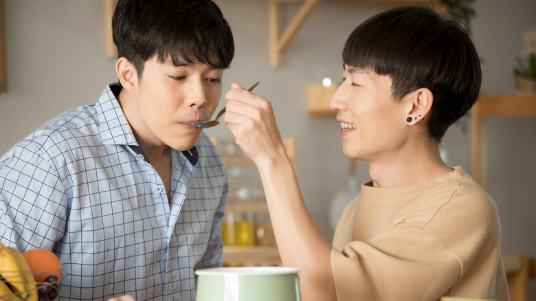 Happy male Asian couple feed each other soup from a spoon in modern kitchen