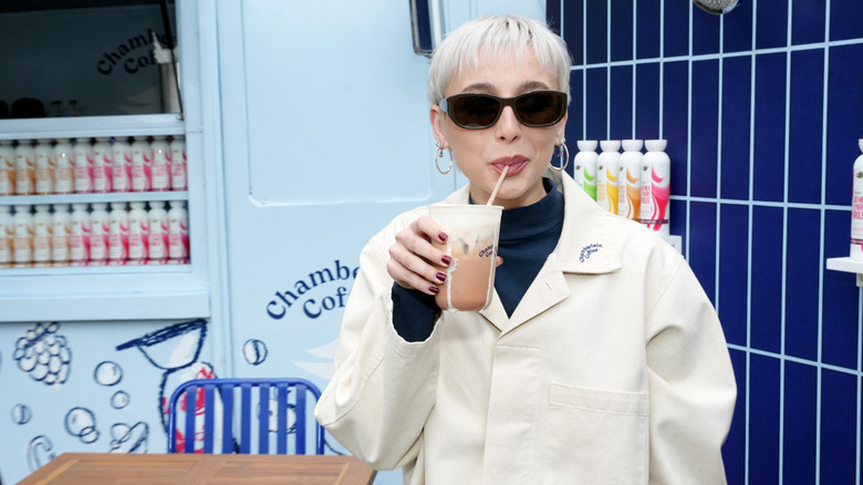 Emma Chamberlain in a white jacket and sunglasses drinking out of a straw in front of a blue and white tiled wall and a Chamberlain Coffee truck