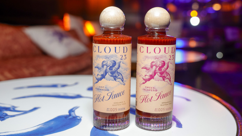 Two bottles of Cloud 23 salsa in decorative glass bottles with round wooden stoppers on a white cafe table with blue peppers painted on it