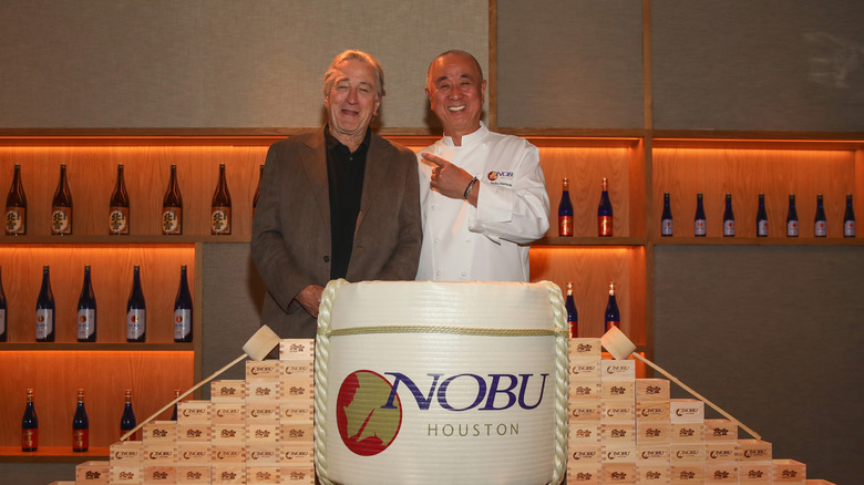 Chef Nobu Matsuhisa wearing a white chef's jacket and pointing at a smiling De Nire while the two stand in front of a display of saki