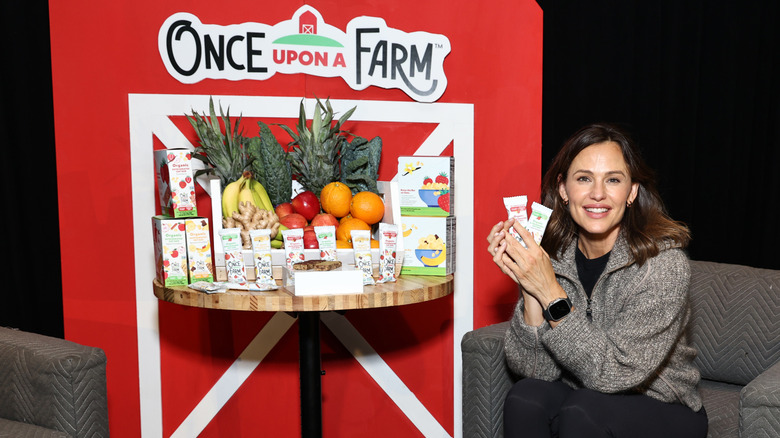 Jennifer Garner smiling and posing with a table of Once Upon a Farm products in front of a miniature red and white barn