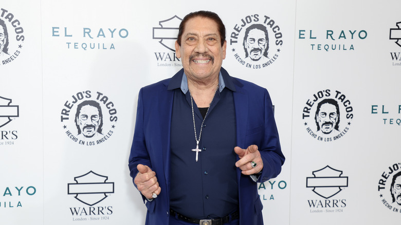 Danny Trejo in a blue shirt and blue jacket with a cross necklace in front of a Trejo's Tacos advertisement board