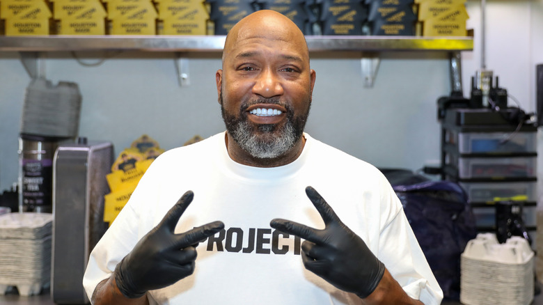 Bun B smiling at the camera while wearing black rubber gloves in an industrial kitchen