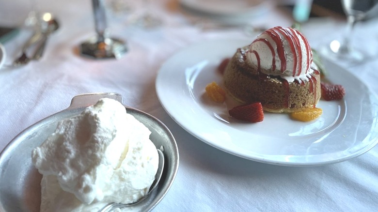 An ice-cream covered, individual-sized cake placed on a white restaurant plate next to a bowl of cream.