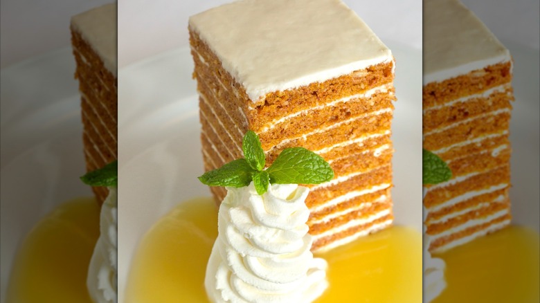 A tall carrot cake, yellow syrup, and whipped cream on a plate.