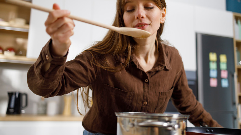 woman tasting food from long wooden spoon while cooking over stovetop with metal pot