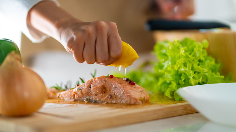woman squeezing lemon over salmon on wooden cutting board with fresh lettuce and whole onion