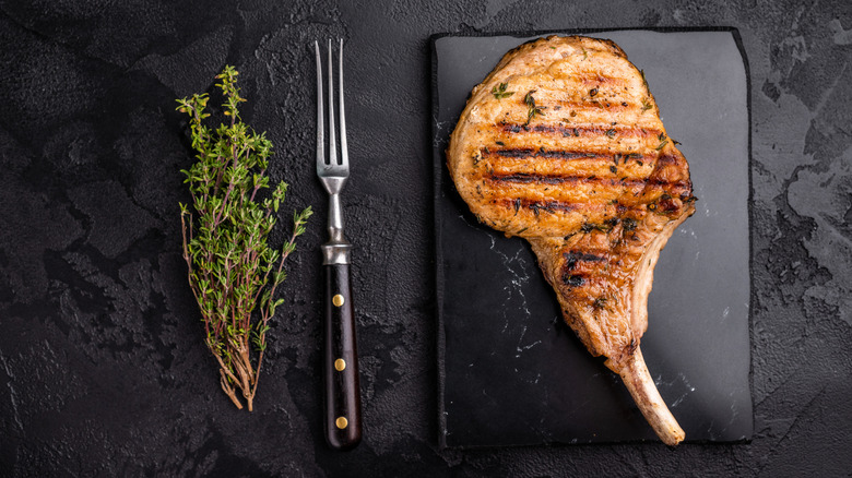 seared pork chop on black tray next to long fork and fresh sprig of thyme