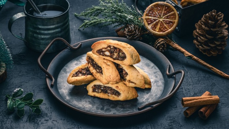 a plate of cuccidati, italian fig cookies, surrounded by a filled mug and pine cones, cinnamon sticks, and leaves