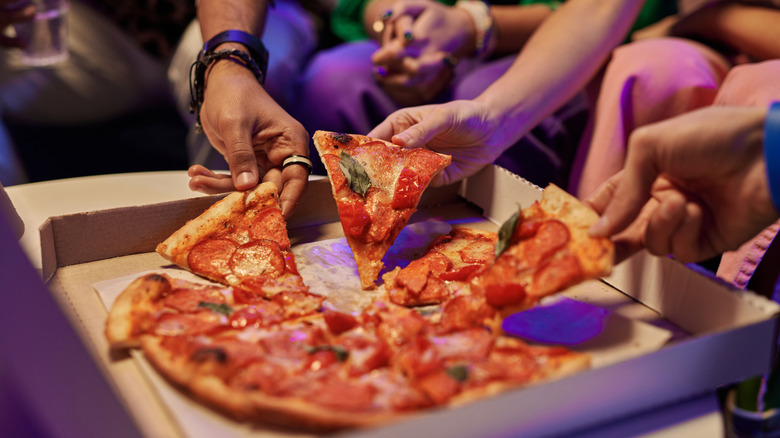 Close-up of slices of appetizing pizza in square cardboard box and hands of young friends taking them and eating while enjoying home party