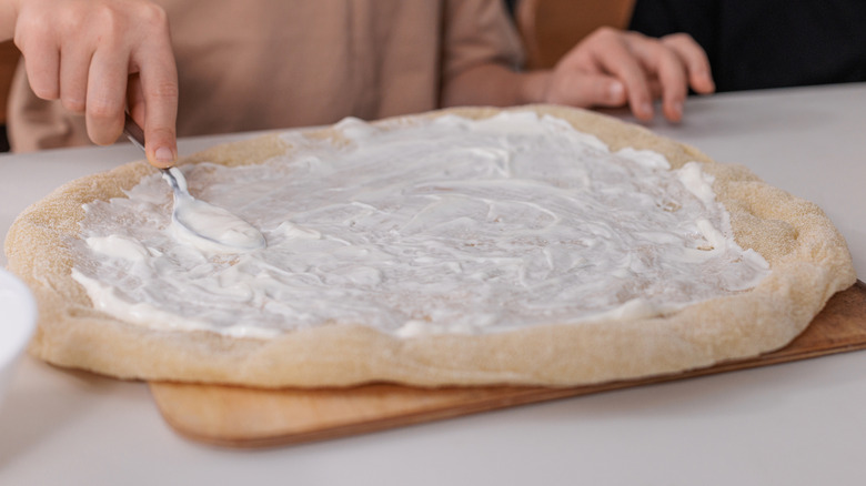 Children preparing pizza at home and spreading white sauce on dough,