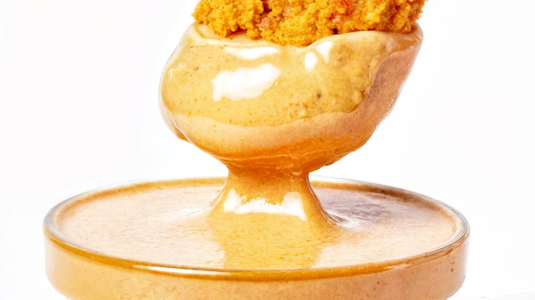 A hand dips a crispy chicken strip into a clear glass cup of honey mustard sauce. The golden chicken is partially covered in rich, creamy dip.