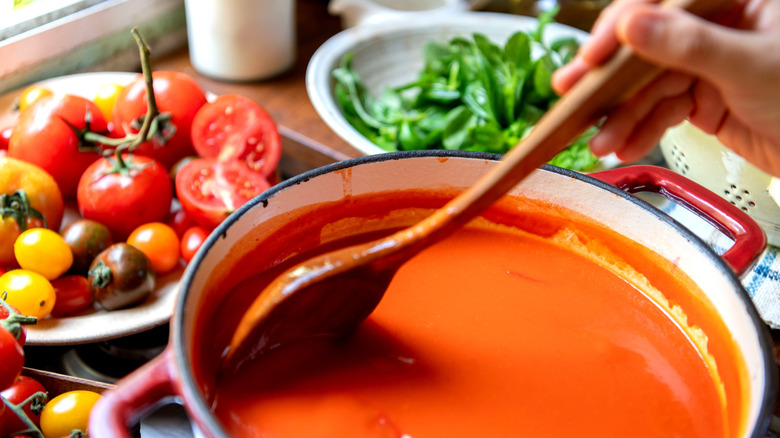 Hand stirring tomato soup with fresh greens and tomatoes in background