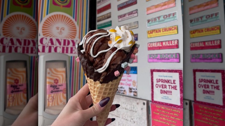 Ghirardelli Squared Chocolate Ice Cream in a dipped waffle cone with whipped cream and lucky charms marshmallows in front of a wall of flavor labels