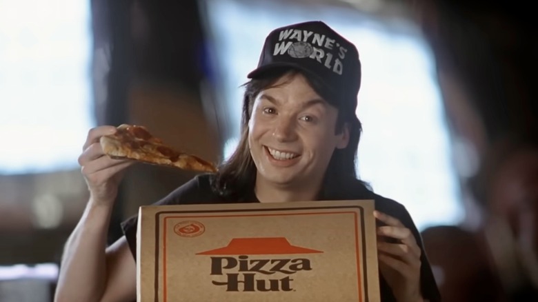 Wayne Campbell holds up a slice of Pizza Hut pizza, with a smile