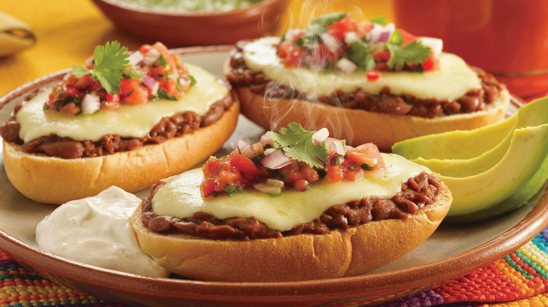 molletes with beans, cheese, and pico de gallo on a beige and brown plate garnished with sliced avocado and sour cream