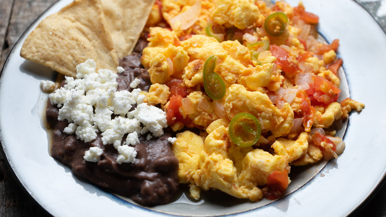 Mexican-style scrambled eggs with green chiles on a white plate with refried beans, cotija cheese, and tortilla wedges