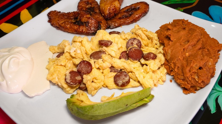 scrambled eggs with sliced sausage on a square white plate with refried beans, fried plantains, sliced avocado, and sour cream