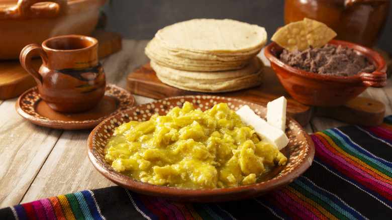 a plate of scrambled eggs with green salsa shown with cheese, corn tortillas, and refried beans pictured in a Mexican-style table setting