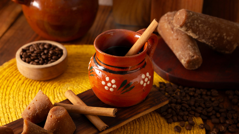 cafe de olla in a brown patterned mug shown with piloncillo, cinnamon sticks, and coffee beans