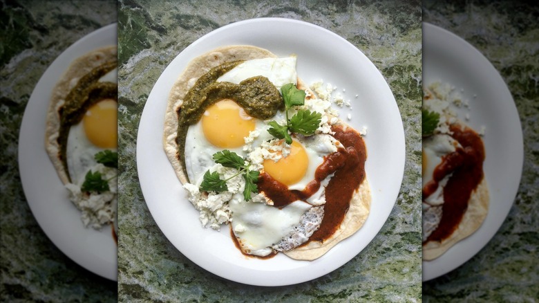 two fried eggs on two tortillas, one topped with green salsa and the other with red salsa, both on a white plate
