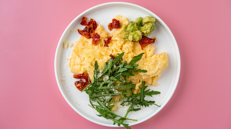 Scrambled eggs with sun dried tomatoes, avocado, and arugula on a pink background