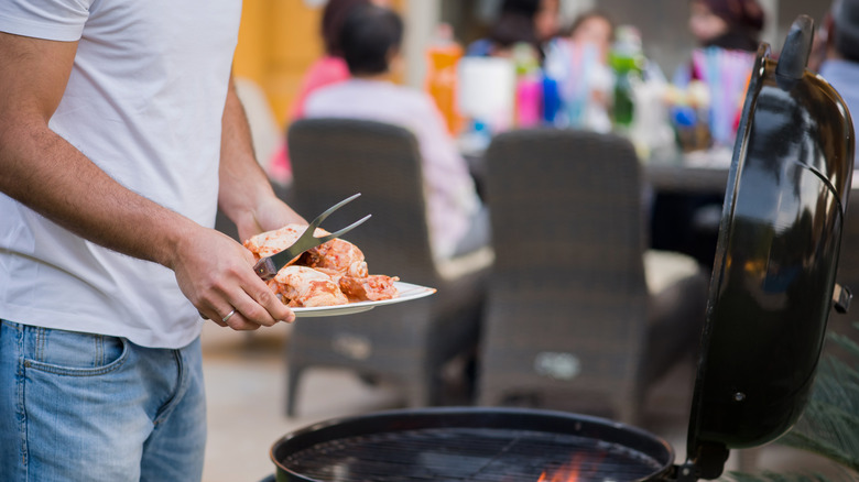 person cooking meat on a grill while people in the background sit at a picnic table