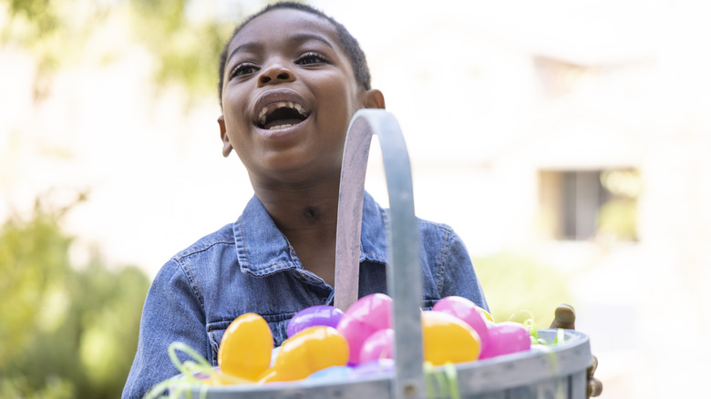 A young boy carrying a large Easter basket full of plastic eggs, smiling excitedly off to the side