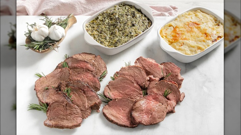Slices of Rastelli's beef tenderloin on butcher paper with casserole dishes of sides and two head of garlic in a small basket.