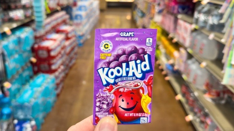A hand holding a packet of grape Kool-Aid mix.