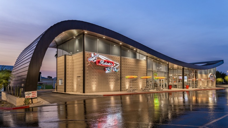 An art-deco style roadside diner location of Roberto's Taco Shop with a neon sign and colorful umbrellas on the outside tables.