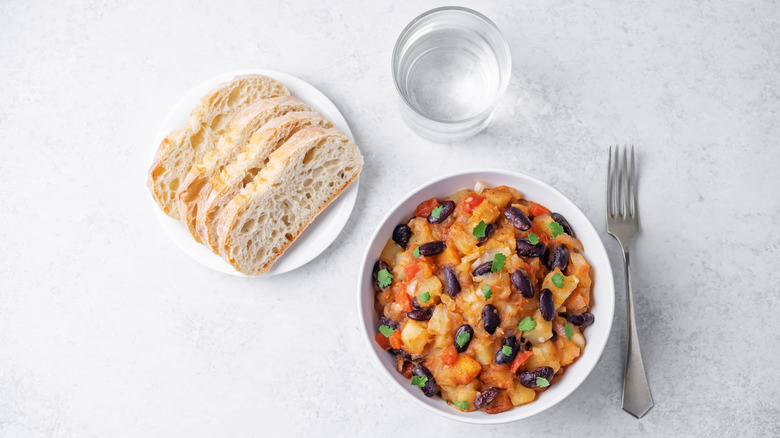 A bowl of bean stew sits on a white table with a plate of bread to the side, a glass of water and a fork