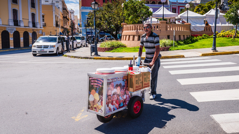 Man crosses a street in Puerto Rico pushing an ice cream cart.