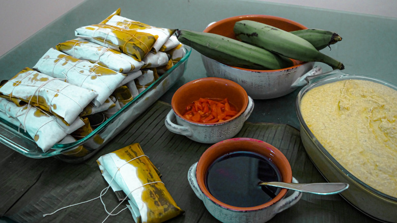 Puerto Rican pasteles wrapped in banana leaves next to bowl of green plantains, bowl of mashed banana, bowl of broth, and bowl of red veggies