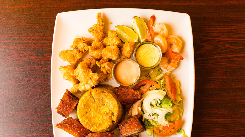 A mound of mashed plantains on plate with sauces, fried pork, fried chicken, shrimp, and fresh salad