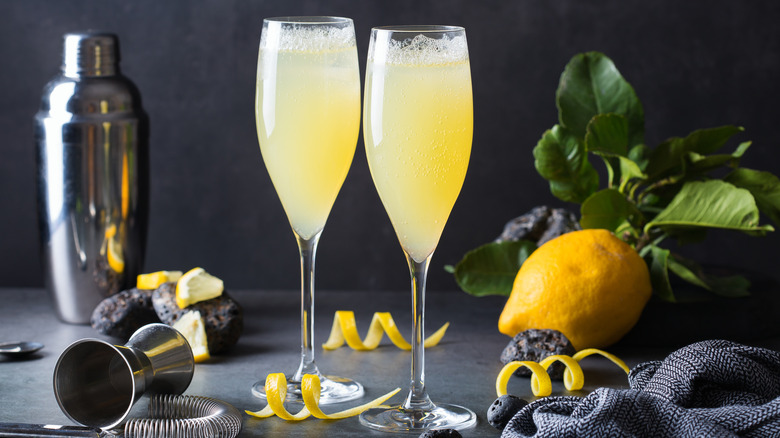 A pair of Champagne flutes filled with pale yellow drink are on a counter next to a lemon and a cocktail shaker.