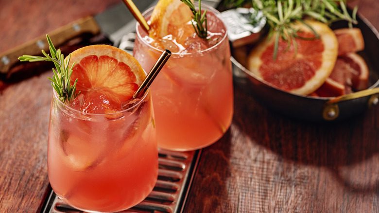 A pinkish colored Paloma cocktail garnished with a lemon wheel and sprig of rosemary.